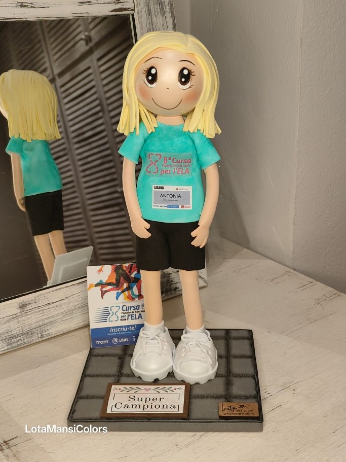 Muñeca de pelo rubio con camiseta azul turquesa, pantalones negros y zapatillas blancas, participando en una carrera a beneficio del ELA