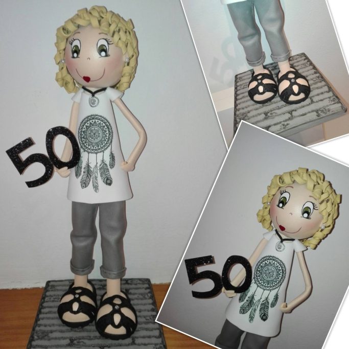Muñeca con cabello rizado, camiseta blanca con un diseño, pantalones grises y sandalias negras, sosteniendo un 50. Los años que cumple.