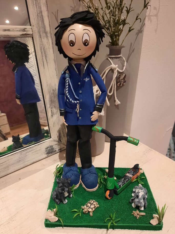 Figura de un niño de pie, con chaqueta azul, patinete y elementos decorativos en la base.