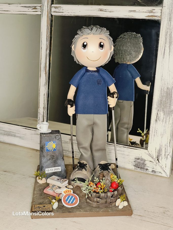 Muñeco con palos de caminar , vestido con camiseta azul y pantalones grises, con piedra del camino, libros, una cesta con verduras y un café