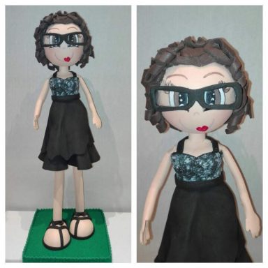 Figurita de goma eva con vestido negro, gafas y cabello rizado. Base verde.