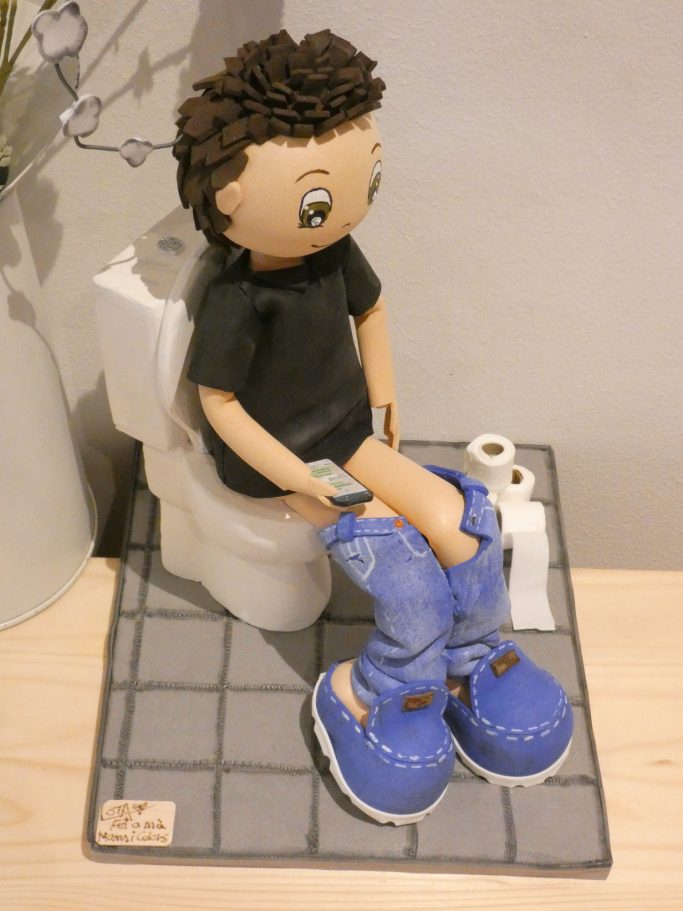 Muñeco sentado en un inodoro con pantalones bajados, zapatillas azules y leyendo whatsApp. Rollos de papel de wc en el suelo