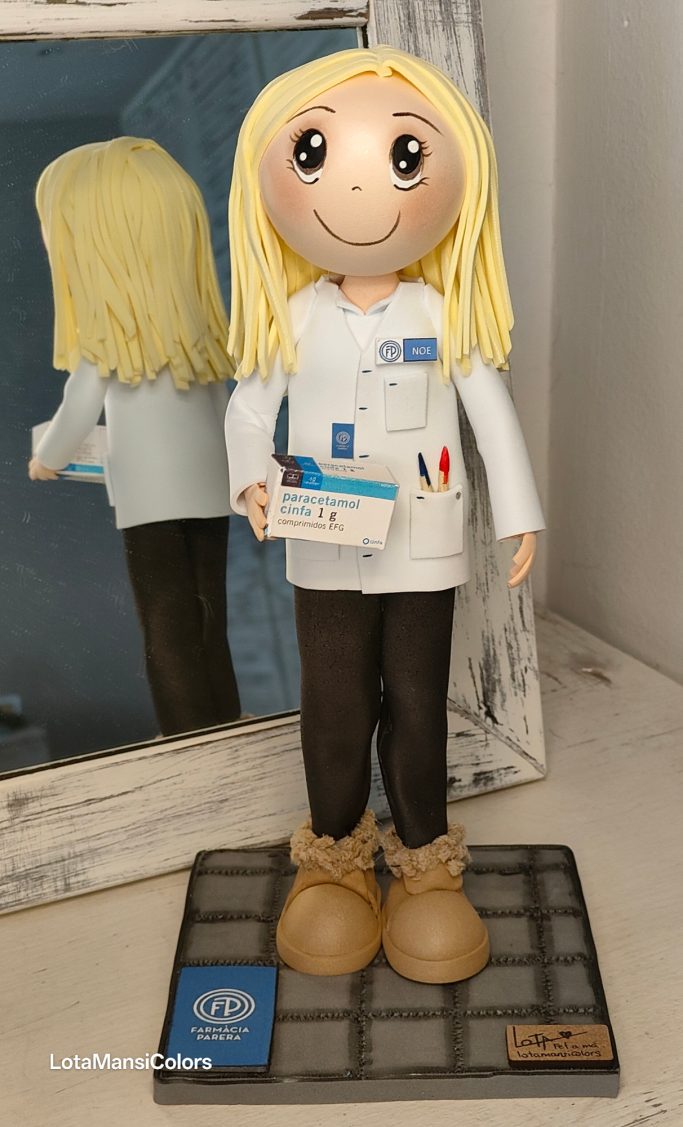 Muñeca de cabello rubio, vestida con bata y pantalones, en sus manos  una caja de paracetamol y en el bolsillo lleva unos bolígrafos.