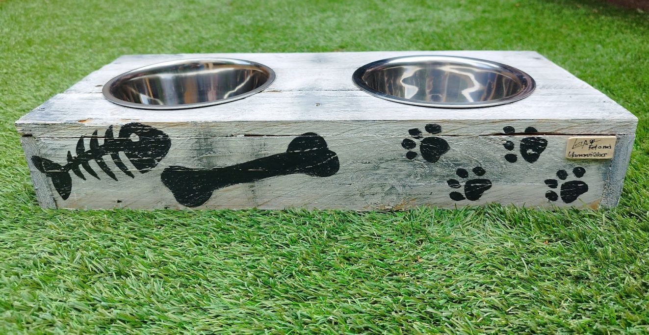 Comedero doble para mascotas con dibujos de huesos y huellas en un fondo de césped.