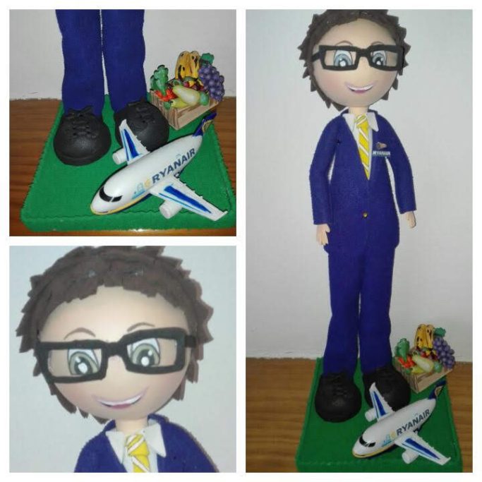 Muñeco de un piloto con traje azul, gafas, junto a una maqueta de avión y una caja con frutas