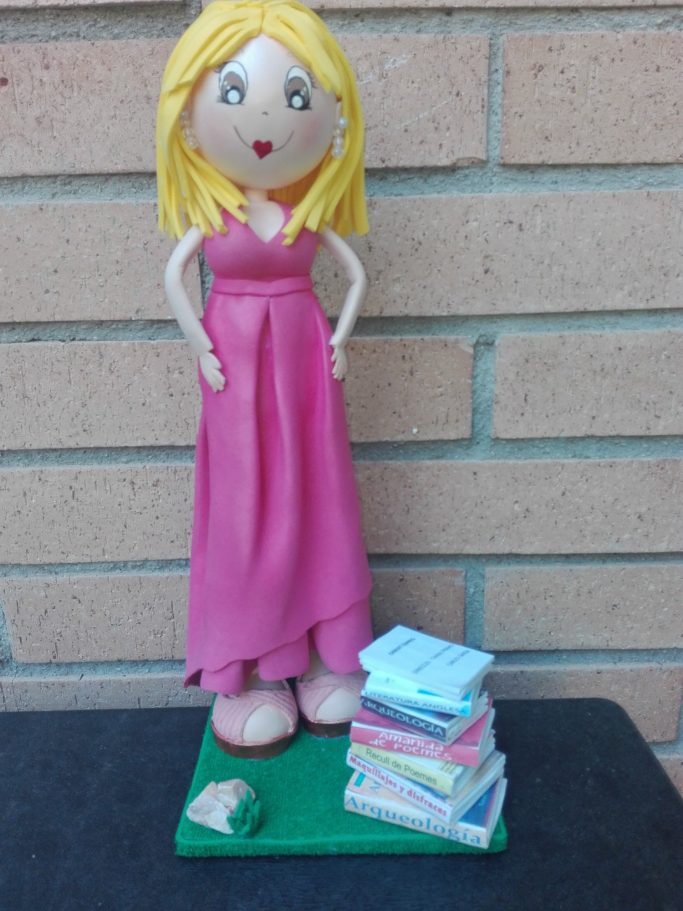 Muñeca de mujer con vestido rosa junto a una pila de libros sobre césped.