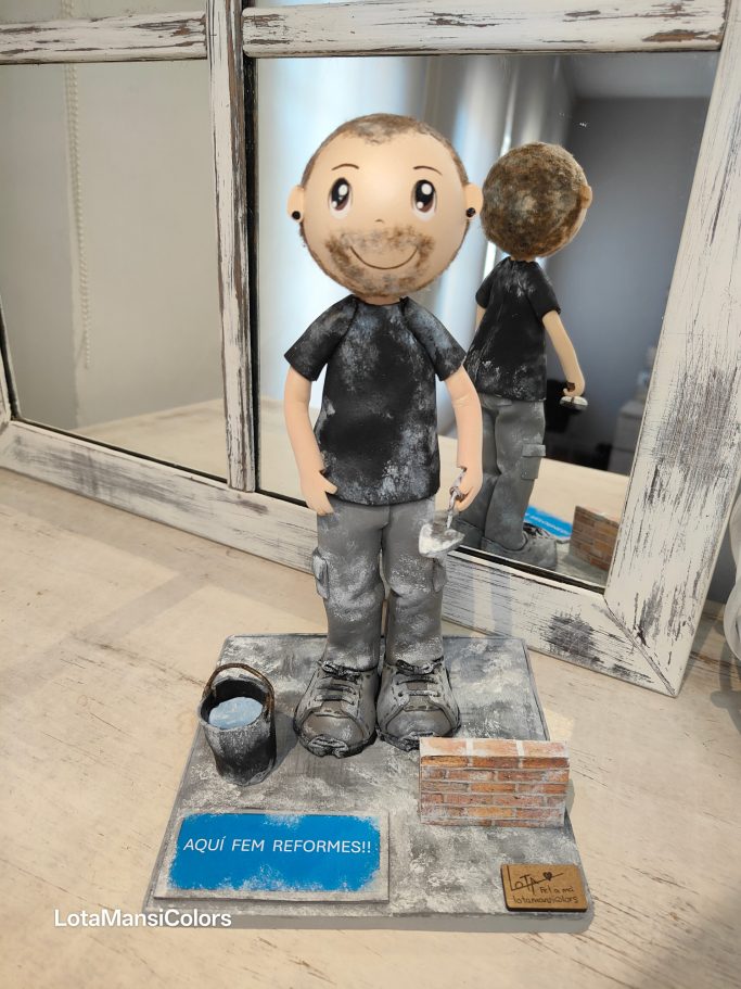 Fofucha de hombre albañil, sucio de trabajar, lleva en la mano una paleta y en el suelo un cubo de agua con una pared de ladrillos.