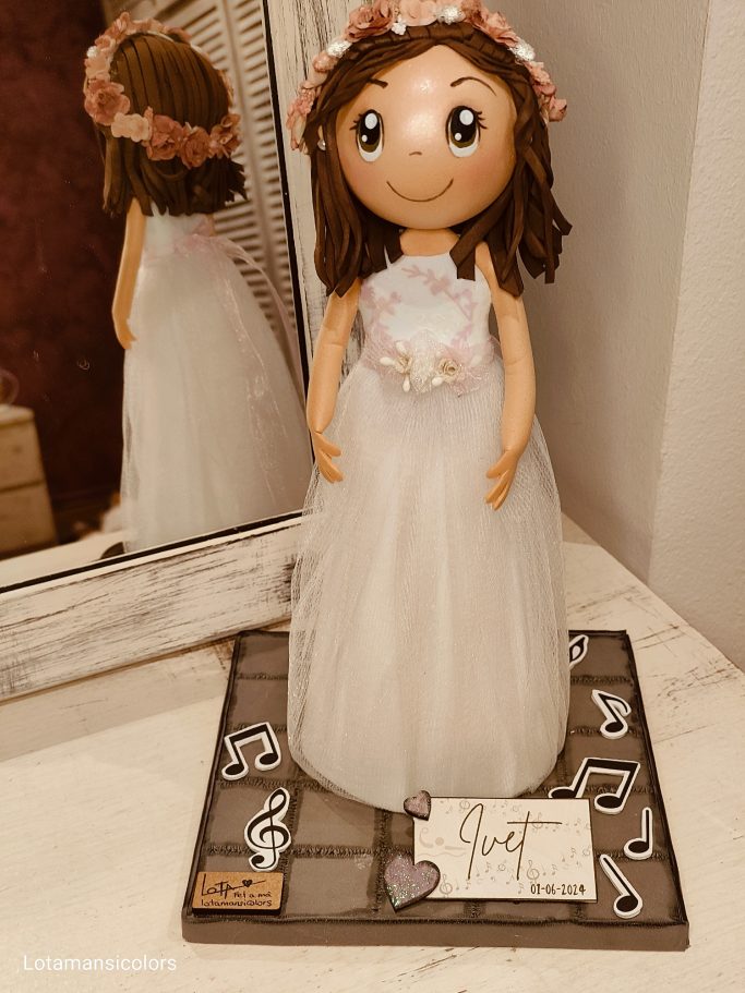 Muñeca de comunión con vestido blanco y corona de flores, sobre una base decorativa.