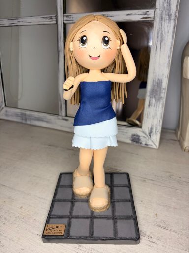 Fofucha de una niña de pie, con cabello largo y rubio, vestida con una blusa azul y falda blanca.