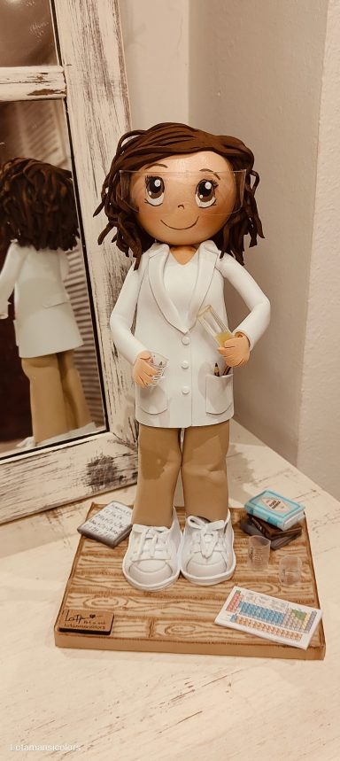 Focucha personalizada, con cabello rizado, vestida con bata blanca y pantalones marrones, con una probeta en la mano, y gafas de protección.