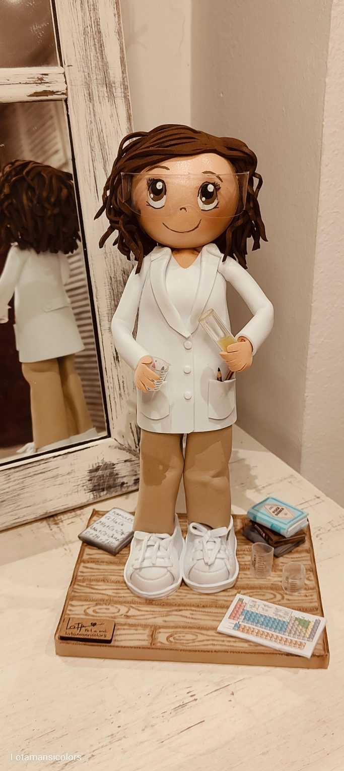 Muñeca con cabello rizado, vestida con bata blanca y pantalones marrones, con una proveta en la mano, y gafas de protección