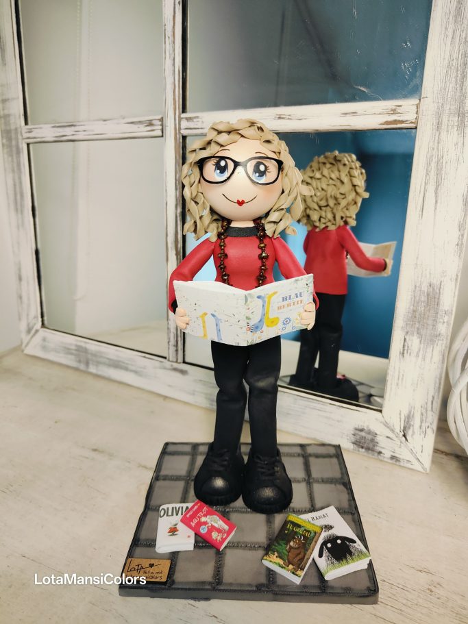 Figura de una mujer de cabello rizado y gafas leyendo un cuento y en el suela más cuentos.